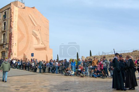 Foto de Tarragona, España - 22 de abril de 2024: Una multitud se reúne para presenciar una procesión de Semana Santa en Tarragona con individuos en atuendo tradicional en medio de un ambiente solemne. - Imagen libre de derechos