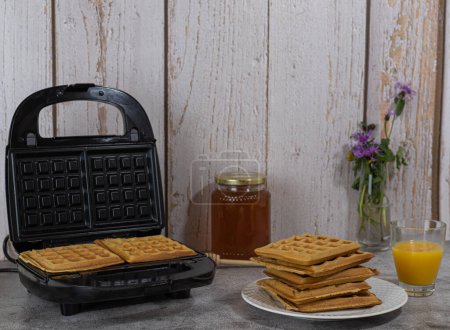 delicious waffles with honey and orange juice. Black waffle maker with fresh waffles. aged white wood background.
