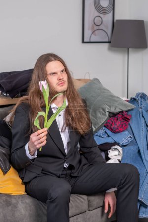 Photo for Confused, distracted and depressed man with long, brow hair looking down at something and holding a pink tuplip. Man wearing a black suit is sitting on the couch in total mess. - Royalty Free Image