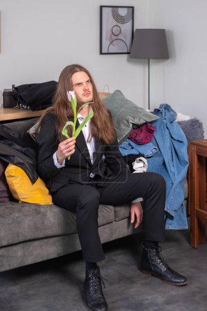 Photo for Confused, distracted and depressed man with long, brow hair holding a pink tuplip. Man wearing a black suit is sitting on the couch in total mess. - Royalty Free Image