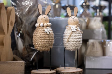 Cute smiling Easter bunnies made of cotton thread with wooden ears and heads on a wooden base on a shelf. Wooden Easter bunny in a box, blurred wrapped decoration materials in the background.