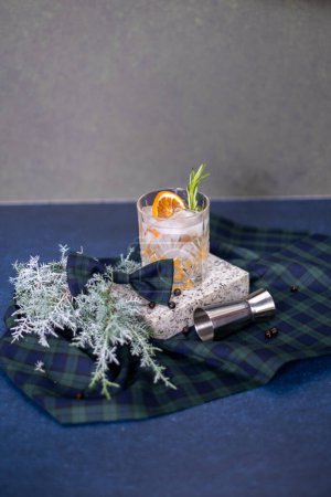 Photo for Crystal glass with gin and tonic cocktail with orange slice, rosemary branch and ice, checkered bow, alcohol measuring cup and juniper branch in the background, Christmas mood. Copy space - Royalty Free Image