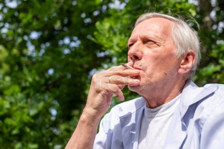 Ein alter Mann mit einer Zigarette im Park nimmt einen Schluck Zigarettenrauch und reflektiert über das Leben, perfekt für Erzählungen über das Altern und persönliche Entscheidungen, ungesunde Lebensstile, Rauchen, 
