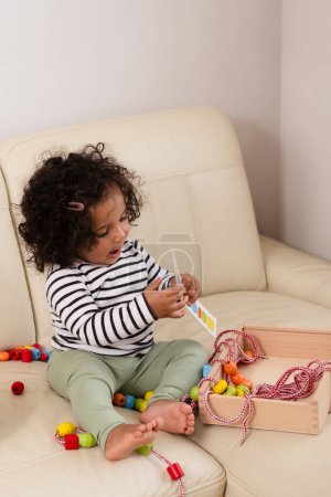 A mixed race toddler with curly hair plays with colorful toys on a cream sofa, immersed in playtime. Ideal for childhood and learning themes. High quality photo