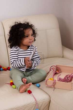Smiling African American toddler with tousled curls plays with beads on a neutral couch, embodying innocence and engagement. High quality photo