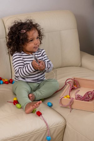 Curious mixed-race child with curly hair sits on a sofa, exploring a wooden toy. Perfect for themes of diversity and childhood curiosity. High quality photo