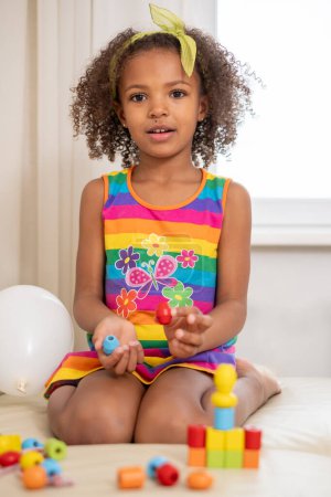 Young mixed race girl playing with colorful blocks, curly hair, yellow bow, rainbow dress. A bright, playful scene ideal for educational themes, and learning through playing. High quality photo