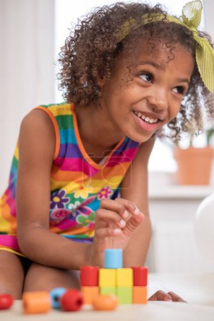 Sonriente chica afroamericana de pelo rizado con sonrisas de lazo amarillo mientras apilan bloques, vestido de rayas de arco iris vibrante. para el contenido de la alegría en el aprendizaje y el juego, la terapia de juguetes..