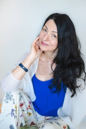 Besinnliche Frau Ende 40, mit einem sanften Lächeln, in blau gekleidet. Erfasst die emotionale Komplexität des Eintritts in die Wechseljahre, emotionale Herausforderungen und Midlife-Crisis. Hochwertiges Foto