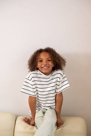 A joyful mixed-race child with a bright smile and curly hair, dressed in a striped shirt, sits comfortably on a beige couch against a white background, isolated. High quality photo
