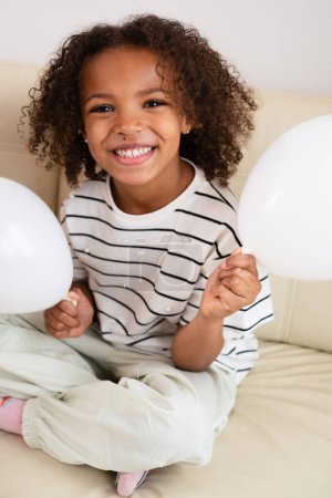 A young African American girl with a bright smile and curly hair holds white balloons, laughing and joyful of sibling relationships, and National Siblings day.. High quality photo