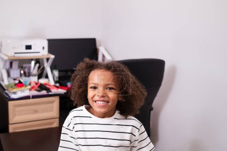 Ein nettes und entzückendes schwarzes Mädchen mit lockigem Haar lächelt auf einem Schreibtisch im Hintergrund. Themen der zu Hause eingeschulten Kinder, des Lernens und des Nationalen Geschwistertages. Hochwertiges Foto