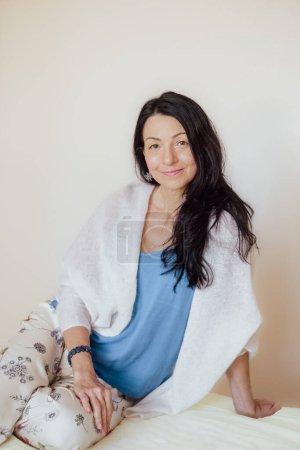 Photo for Smiling woman with black hair, blue top, white shawl, sitting comfortably. Her relaxed posture and warm expression evoke a sense of homey contentment. High quality photo - Royalty Free Image