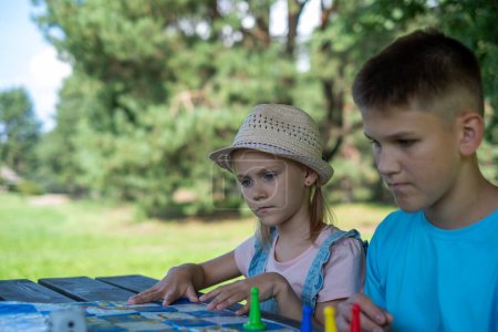 A boy in a blue shirt and a girl in a straw hat focused on a board game in a nature setting. This image perfectly captures childhood, leisure, and learning, National siblings day. High quality photo