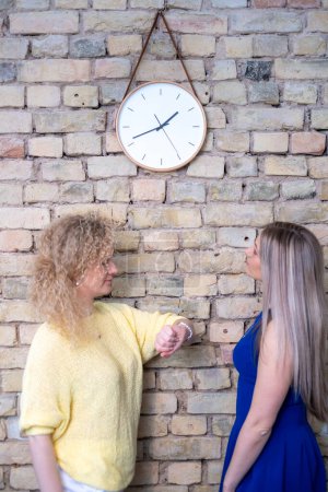 Zwei Frauen, eine in Gelb und die andere in Blau, schauen unter einer Wanduhr auf ihre Uhren und beschwören Zeitmanagement und Dringlichkeit für Themen wie Unternehmens- und Wachstumsberatung herauf. Hochwertiges Foto