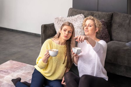 A woman in a yellow sweater listens as her friend in white points and explains, tea cups in hand, in a home setting, a talking, therapy session . High quality photo