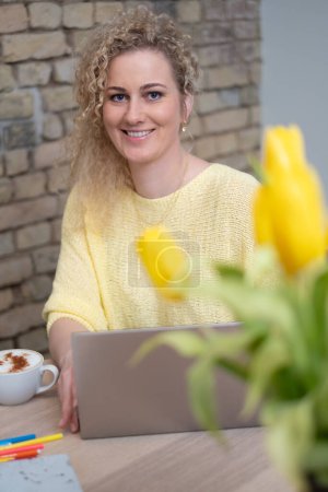 Eine fröhliche Frau im gelben Pullover arbeitet an ihrem Laptop, ihr Lächeln vermittelt Zuversicht und Positivität, während gelbe Tulpen ihren optimistischen Ausblick auf das Wirtschaftswachstum widerspiegeln. Hochwertiges Foto