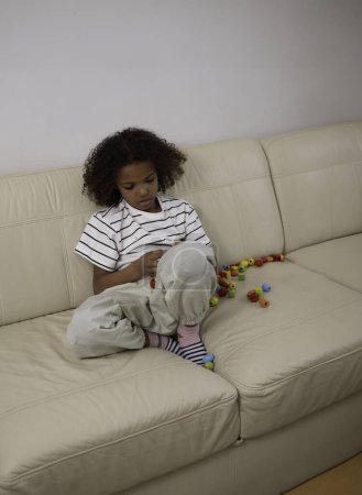 A young mixed-race girl focused on sewing a cloth doll on a cream couch, surrounded by colorful beads. playing and learning, for themes of child development and eco-friendly toys. High quality photo