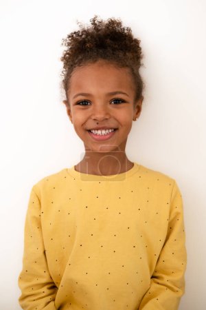 A cheerful young african american girl in a dotted yellow sweater stands against a white background. Her smile exudes warmth and innocence, ideal for themes of joy and childhood. High quality photo