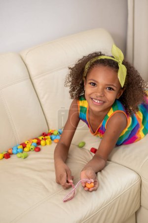 Cheerful curly-haired mixed race girl wearing a rainbow dress, playing with beads, radiates happiness and the simplicity of childhood joy in a cozy home setting. High quality photo