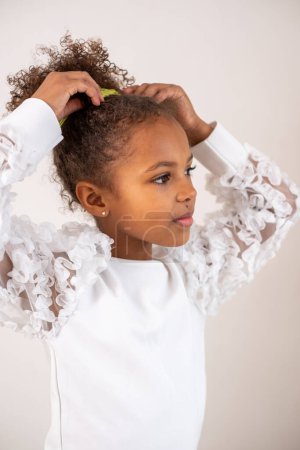 A contemplative mixed race girl with curly hair, wearing a white frilly dress, looks to the side against a neutral backdrop, evoking curiosity and elegance. High quality photo