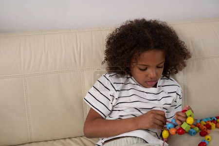 Focused African American young girl with curly hair, engaging with colorful beads, exemplifying playful learning and child development in a homely setting. High quality photo