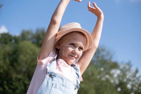 Ein fröhliches kleines Mädchen mit Strohhut und Jeanshose streckt die Arme gegen den blauen Himmel und strahlt Glück und Freiheit aus. im Zusammenhang mit Sommer, Spielzeit und Kindheit. Hochwertiges Foto