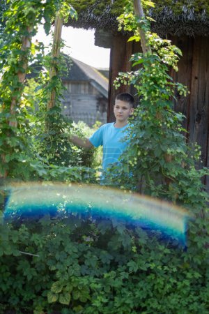 Ein Junge im blauen T-Shirt steht inmitten sattgrünen Laubes in einem rustikalen Garten, sein Gesichtsausdruck ist nachdenklich und ruhig. Perfekt, um das Engagement der Jugend für Natur und ländliches Leben zu veranschaulichen. Hochwertiges Foto
