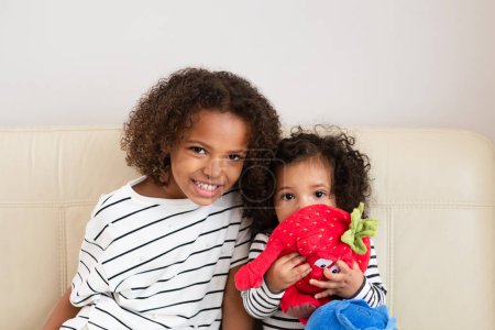 Two smiling mixed race siblings embracing, the older with a striped shirt, the younger holding stuffed toys, portray warmth and family bonding, and national siblings day. High quality photo