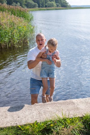 A man lifts a giggling child by the lake a joyous, sunny day. Captures family fun, summer activities, used for health insurance for active summers. High quality photo