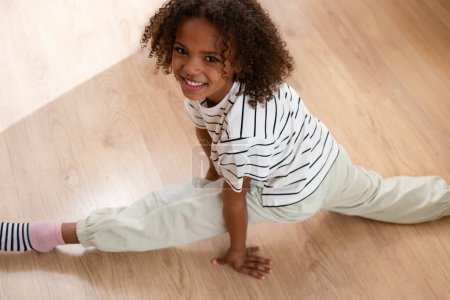 Téléchargez les photos : Un enfant souriant de race mixte portant une chemise rayée faisant une fente ludique sur un plancher de bois, incarnant la joie insouciante de l'enfance et l'athlétisme. Photo de haute qualité - en image libre de droit