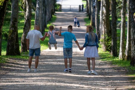 A father playfully swings a child between him and her siblings, as they walk hand in hand down a tree-lined path, capturing the essence of family bonding and shared joy. High quality photo