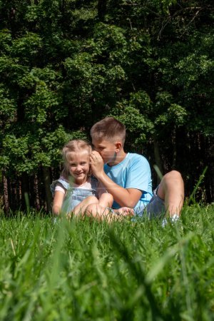 The boy in casual attire leans close, whispering to the girl in denim, a tender sibling scene set against the park's verdant backdrop, National siblings day and happy friendships. High quality photo