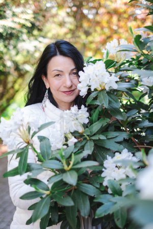 A joyful middle-aged woman peers through blooming white rhododendrons, her smile reflecting a sense of renewal and connection with nature, therapy in the spring. High quality photo