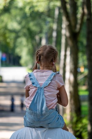Ein junges Mädchen wird auf den Schultern ihres Vaters getragen und genießt einen sonnigen Tag in einem von Bäumen gesäumten Park, eine Momentaufnahme der Unschuld und der einfachen Freuden der Kindheit. Hochwertiges Foto