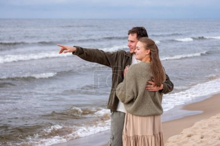 Man pointing to the horizon, embracing woman from behind, both looking sea-ward. A perfect portrayal of shared discovery and wanderlust, or weeding celebration themes. High quality photo