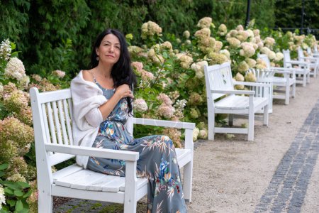 Femme dispendieuse dans un salon de robe florale sur un banc de jardin blanc, son châle drapé avec désinvolture, au milieu d'une voie d'hortensias luxuriantes, évoquant un moment de solitude paisible. Photo de haute qualité