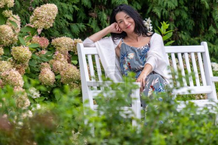 En un exuberante jardín, una mujer con una sonrisa contenta descansa en un banco, envuelto en vegetación y los suaves tonos de hortensias, una imagen de ocio sereno. Foto de alta calidad
