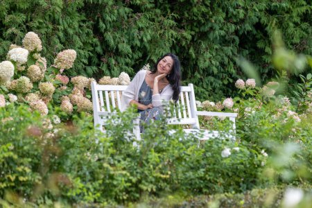 Auf einer Parkbank liegend, verkörpert eine Frau mit zufriedenem Lächeln, umgeben von dichten Hortensien, entspannte Eleganz in der Umarmung der Natur. Hochwertiges Foto