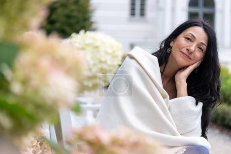 A woman wrapped in a cream shawl sits amidst blooms, her content smile reflecting self-acceptance and grace at midlife amidst hormonal transition. High quality photo