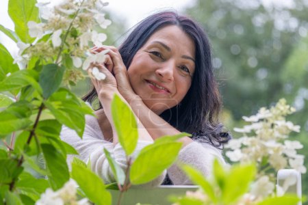Eine lächelnde Frau mittleren Alters in einem grünen Blumengarten drückt Glück und Freude über hormonelle Veränderungen und Kämpfe in der Lebensmitte aus. Hochwertiges Foto