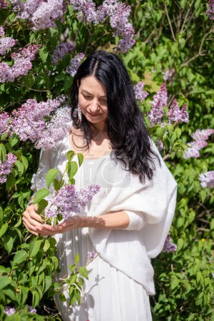 Contemplative woman with dark hair admiring lilac blooms, dressed in white, surrounded by lush greenery, a moment of connection with nature. High quality photo
