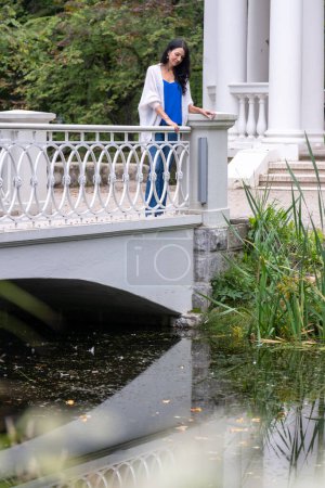 Eine Frau lehnt an einer weißen Brüstung an einem Teich, inmitten der Natur, und spiegelt Gelassenheit und Besinnlichkeit wider, was Themen wie Lebensmitte und Ruhe suggeriert. Hochwertiges Foto