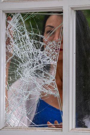 Eine Frau blickt durch ein zersplittertes Fenster, ihr Blick wird von einem Netz aus Rissen durchtrennt, das eine eindringliche Metapher für Not oder Zerrüttung schafft. Hochwertiges Foto