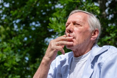 Un anciano disfruta de un cigarrillo al aire libre, con un fondo de exuberante vegetación, capturando un momento personal y reflexivo. Foto de alta calidad