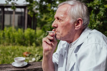 A contemplative older man smokes leisurely, overshadowed by verdant foliage, capturing a private moment of solitude. High quality photo
