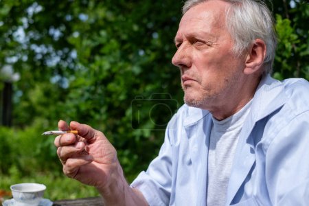 Tief in Gedanken pausiert ein älterer Herr für eine Raucherpause im Freien, inmitten der Frische eines lebendigen Gartens, der im hohen Alter süchtig ist. Hochwertiges Foto