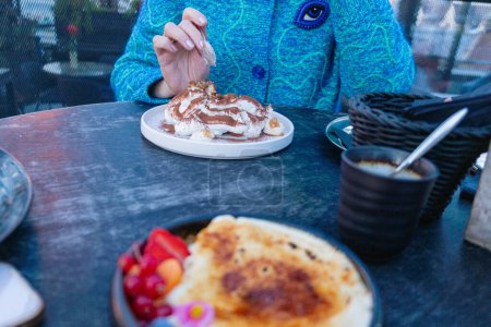 Eine Person genießt einen Teller Tiramisu im blauen Pullover in einem Café im Freien, mit Kaffee und frischem Obst an der Seite. Hochwertiges Foto