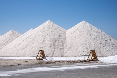 Pyramid like bright white salt heaps dominate the landscape, while wooden trestles add a rustic touch to the salt extraction site on a blue background, copy space.. High quality photo