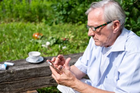 Senior man in glasses focused on his smartphone at a rustic wooden table outdoors, with a tranquil garden and tea cup in the background. High quality photo